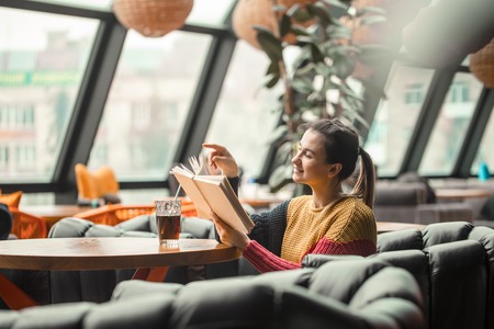 Young beautiful woman in orange sweater reading interesting book in cafe. The concept of readingの写真素材