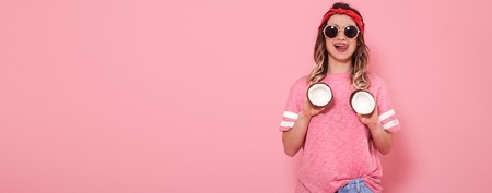 Beautiful young girl in pink t-shirt and glasses holding funny sexy coconuts on pink background. Summer, Rest. Place for text. Advertising conceptの写真素材