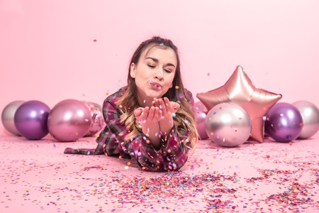 Funny girl blows confetti in her hands. Color Balloons on a pink background. The concept of the party and holidayの写真素材
