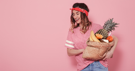 Horizontal portrait of hipster girl in stylish clothing, with bag with fruit, on pink background, lifestyle conceptの写真素材