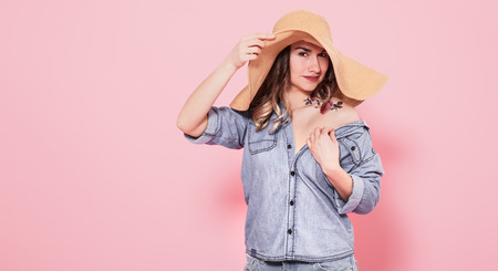 Horizontal portrait of a stylish girl in a summer big hat and denim dress with tattoo on her body, on a pink background, lifestyle conceptの写真素材