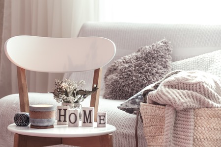 Chic interior for a house. Candles, a vase with flowers with wooden letters of the home on wooden white chair. Sofa and wicker basket with cushions in the background. Home decoration.の写真素材