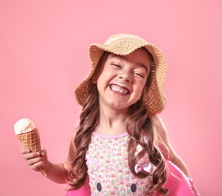 Portrait of a little cheerful girl in a summer hat with ice cream in her hands and a swimming circle, on a colored pink background, the concept of summerの写真素材