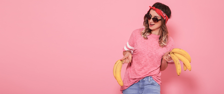 Beautiful young girl in pink t-shirt and glasses holding funny bananas on pink background. Summer, Rest. Place for text. Advertising conceptの写真素材