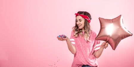 Funny girl in a pink t-shirt with balloons and confetti gives a smile and emotions on a pink background. The concept of the party and holidayの写真素材