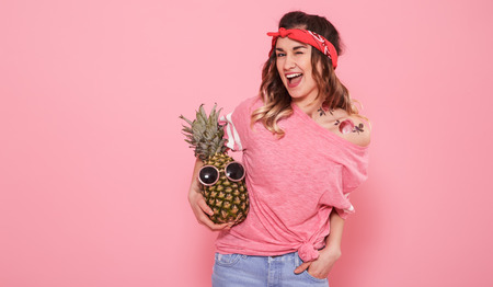 Horizontal portrait of hipster girl with tattoo, and pineapple in sunglasses, on pink background, lifestyle conceptの写真素材