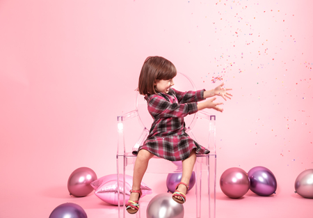 Funny child sitting on a transparent stylish chair on a pink background. Little girl having fun with confetti. The concept of celebration and fun.の写真素材