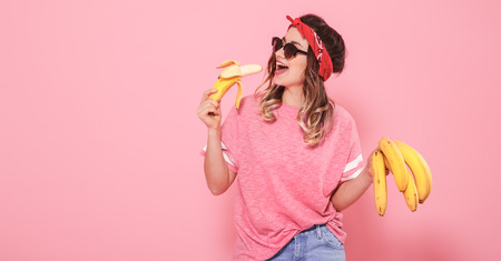 Horizontal portrait of hipster girl in stylish clothes with sunglasses, with bananas in their hands, on a pink background, lifestyle conceptの写真素材