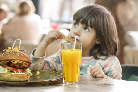 Little cute girl eating a fast food sandwich with fries and orange juice in a cafe. Fast food concept.の写真素材
