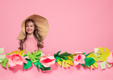 Portrait of a little girl in a summer hat on a pink background with paper flowers, place for text, summer advertising conceptの写真素材
