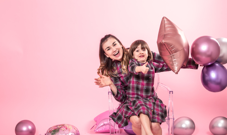 Funny mom and child sitting on a transparent stylish chairs pink background. Little girl and mother having fun with balloons and confetti. The concept of celebration and fun.の写真素材