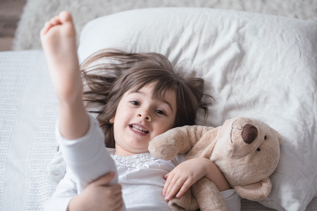Cute little girl sleeps sweetly in a white cozy bed with a soft bear toy, the concept of children's rest and sleepの写真素材