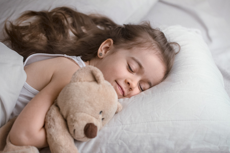 Cute little girl sleeps sweetly in a white cozy bed with a soft bear toy, the concept of children's rest and sleepの写真素材