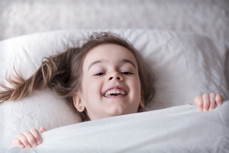 Cute little girl smiling while lying in a cozy white bed with , the concept of children's rest and sleepの写真素材
