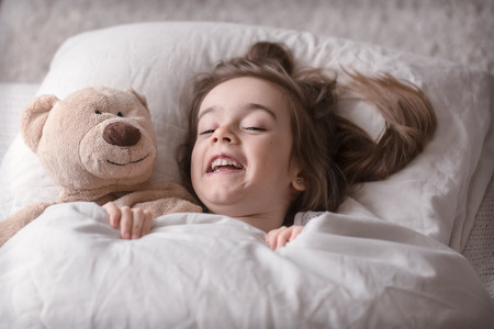 Cute little girl sleeps sweetly in a white cozy bed with a soft bear toy, the concept of children's rest and sleepの写真素材