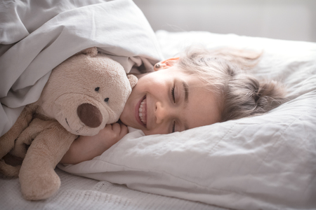 Cute little girl sleeps sweetly in a white cozy bed with a soft bear toy, the concept of children's rest and sleepの写真素材