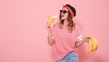 Horizontal portrait of hipster girl in stylish clothes with sunglasses, with bananas in their hands, on a pink background, lifestyle conceptの写真素材