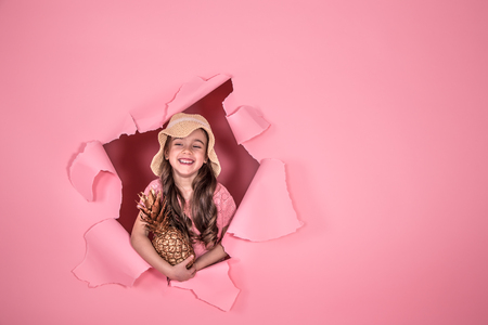 Funny little girl in a beach hat and with pineapple gold color, on a colored pink background, peeking out from the hole in the background, Studio shooting, space for textの写真素材