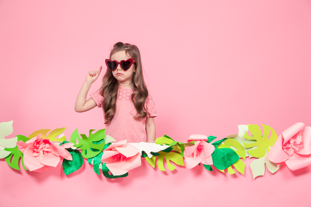 Portrait of little girl with heart shaped sunglasses on pink background with paper flowers, place for text, summer advertising conceptの写真素材