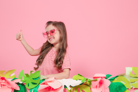 Portrait of little girl with heart shaped sunglasses on pink background with paper flowers, place for text, summer advertising conceptの写真素材