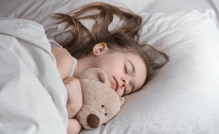 Cute little girl sleeps sweetly in a white cozy bed with a soft bear toy, the concept of children's rest and sleepの写真素材