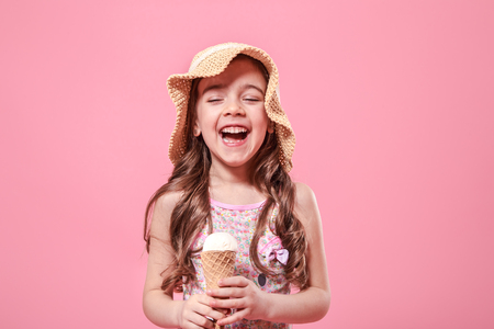 Portrait of a little cheerful girl in a summer hat with ice cream in her hands, on a colored pink background, summer conceptの写真素材