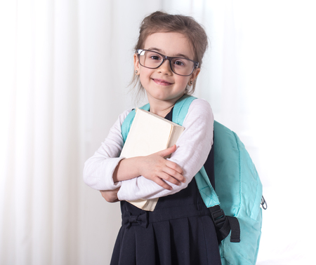 Girl-elementary school student with a backpack and a book with glasses is on a light background .The concept of education and primary schoolの写真素材