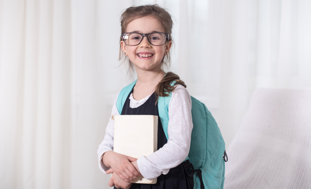 Girl-elementary school student with a backpack and a book with glasses is on a light background .The concept of education and primary schoolの写真素材