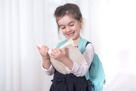 Girl-elementary school student with a backpack on his shoulders reading a book on a light background .The concept of education and primary schoolの写真素材