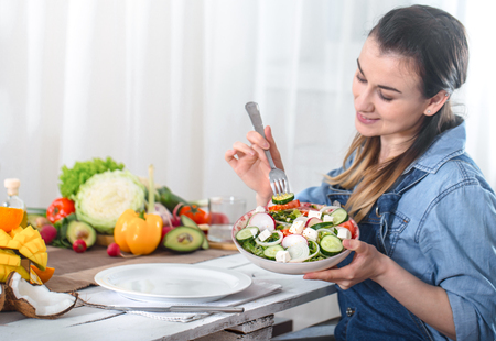 Young and happy woman eating salad with organic vegetables at the table on a light background , in denim clothes. The concept of a healthy home-made food.の写真素材