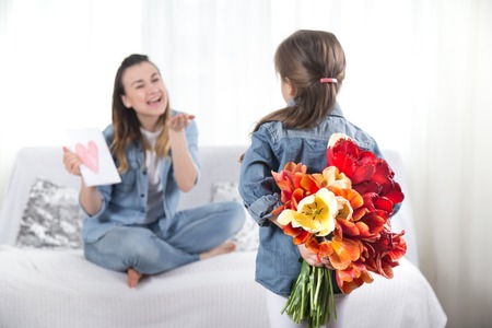Happy mother's Day. Little sweet daughter with a large bouquet of tulips congratulates her mother. In the interior of the living room, the concept of a happy family lifeの写真素材