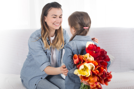 Happy mother's Day. Little sweet daughter with a large bouquet of tulips congratulates her mother. In the interior of the living room, the concept of a happy family lifeの写真素材