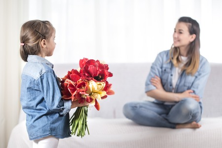 Happy mother's Day. Little sweet daughter with a large bouquet of tulips congratulates her mother. In the interior of the living room, the concept of a happy family lifeの写真素材