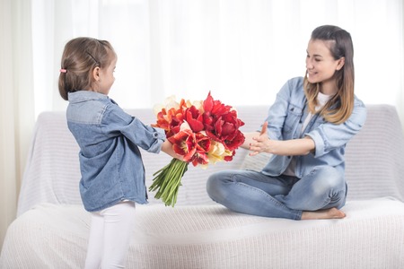 Happy mother's Day. Little sweet daughter with a large bouquet of tulips congratulates her mother. In the interior of the living room, the concept of a happy family lifeの写真素材