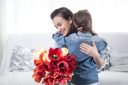 Happy mother's Day. Little cute daughter hugs her mother , and gives a bouquet of tulips, close-up photo. In the interior of the living room, the concept of a happy family lifeの写真素材