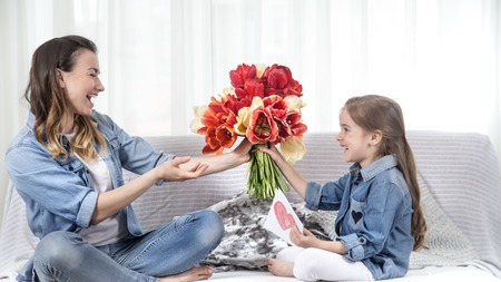 Happy mother's Day. Little sweet daughter with a large bouquet of tulips congratulates her mother. In the interior of the living room, the concept of a happy family lifeの写真素材