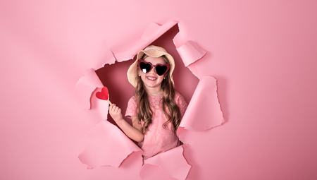 funny little girl looks out from the hole in a beach hat and glasses in the shape of a heart, holding a heart on a stick, on a colored background, a place for text, Studio shootingの写真素材