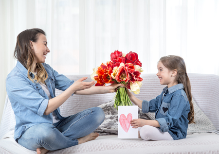 Happy mother's Day. Little sweet daughter with a large bouquet of tulips congratulates her mother. In the interior of the living room, the concept of a happy family lifeの写真素材