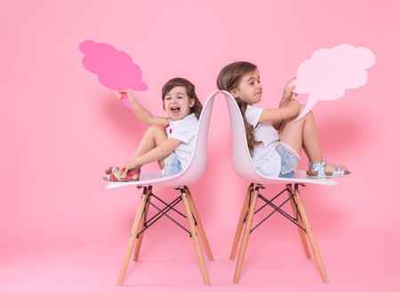 Two little girls on a colored pink background are sitting on chairs with speech icons . The concept of communication and friendshipの写真素材