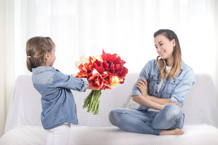 Happy mother's Day. Little sweet daughter with a large bouquet of tulips congratulates her mother. In the interior of the living room, the concept of a happy family lifeの写真素材