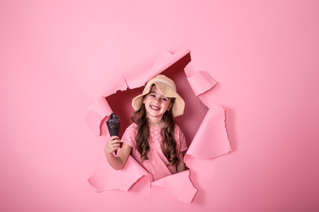 funny little girl peeking out of the hole in a beach hat and ice cream in her hands, on a colored pink background, space for text, Studio shootingの写真素材