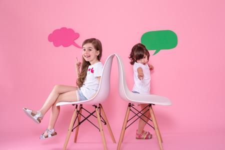 Two little girls on a colored pink background are sitting on chairs with speech icons . The concept of communication and friendshipの写真素材