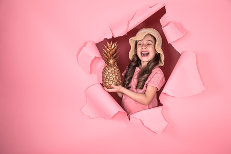 Funny little girl in a beach hat and with pineapple gold color, on a colored pink background, peeking out from the hole in the background, Studio shooting, space for textの写真素材
