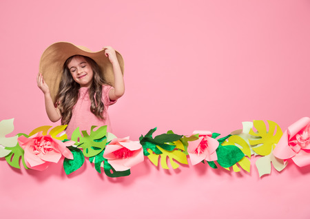 Portrait of a little girl in a summer hat on a pink background with paper flowers, place for text, summer advertising conceptの写真素材