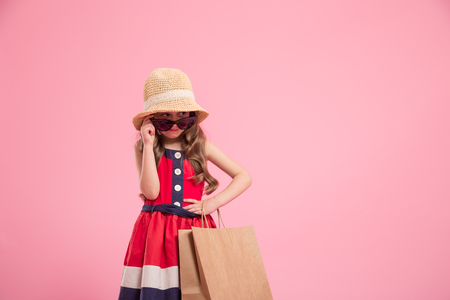 little fashionista with a shopping bag in summer hat and sunglasses , colorful pink background , the concept of children's fashionの写真素材