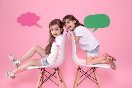 Two little girls on a colored pink background are sitting on chairs with speech icons . The concept of communication and friendshipの写真素材