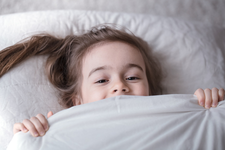 Cute little girl smiling while lying in a cozy white bed with , the concept of children's rest and sleepの写真素材