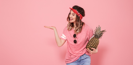 Horizontal portrait of hipster girl in stylish clothes and glasses, with a pineapple in her hands, on a pink background, lifestyle conceptの写真素材