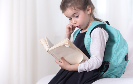 Girl-elementary school student with a backpack on his shoulders reading a book on a light background .The concept of education and primary schoolの写真素材