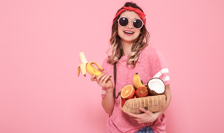 Horizontal portrait of hipster girl in stylish clothes with sunglasses, with healthy food, fruits, on pink background, lifestyle conceptの写真素材